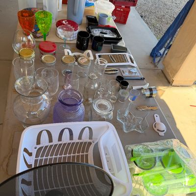 Sale Photo Thumbnail #233: Assorted kitchenware and glassware displayed on an outdoor folding table, including a white plastic dish rack, glass jars, pitchers, and colorful plastic cups.