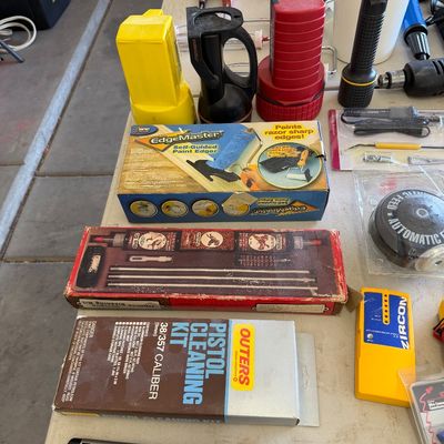 Sale Photo Thumbnail #104: Assorted household and utility items including an Outers pistol cleaning kit, an EdgeMaster paint edger, flashlights, and a Zircon stud finder arranged on a table.