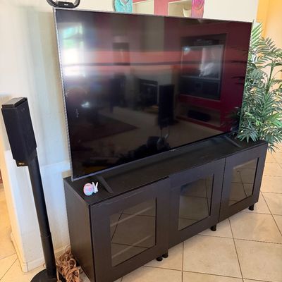 Sale Photo Thumbnail #303: Large flat-screen television on a black three-door media console with glass-front cabinets, accompanied by a tall black speaker stand.