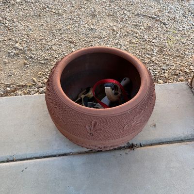 Sale Photo Thumbnail #244: Round terracotta planter with textured, embossed patterns, containing miscellaneous garden tools and a red strap.