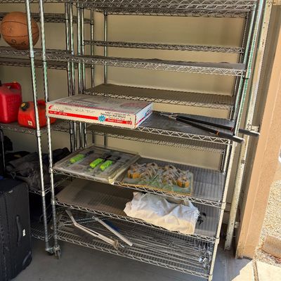Heavy-duty chrome wire shelving unit on casters, holding various garage items including tools, a TV wall mount box, and hardware.