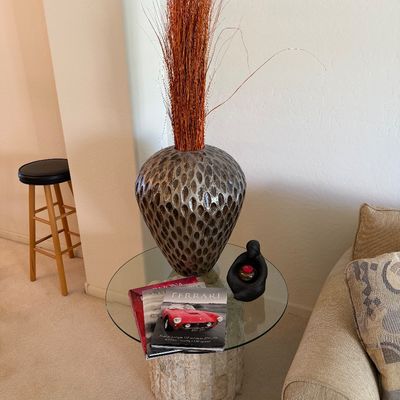 Sale Photo Thumbnail #309: Contemporary glass-top side table with a textured stone pedestal base, styled with a large dimpled metallic vase, decorative branches, and coffee table books.