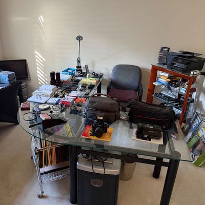 L-shaped glass-top desk with black metal frame, featuring various electronics, camera gear, office supplies, and a paper shredder...