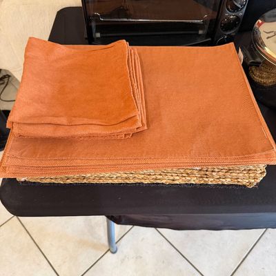 Sale Photo Thumbnail #266: Set of terracotta-colored fabric placemats and matching napkins, stacked on top of woven chargers on a black folding table.