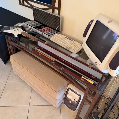 Assorted items on a glass-top console table, including a black electric guitar, a poker chip set in an aluminum case, a pool cue in a...