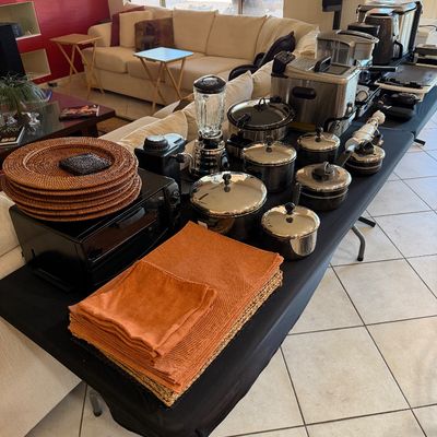 Sale Photo Thumbnail #275: Assorted kitchenware and small appliances displayed on a folding table, including a set of stainless steel pots, a blender, a toaster oven, woven chargers, and orange table linens.