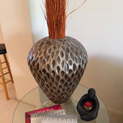 Sale Photo Thumbnail #304: Large textured ceramic vase with a metallic finish, holding dried orange grass, displayed on a glass-top side table alongside books and a small black sculpture.