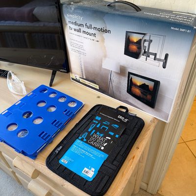 Sale Photo Thumbnail #193: Simplicity medium full-motion TV wall mount in its original box, shown alongside a blue plastic folding board and a black Grid-It accessory organizer.