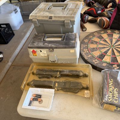 Assorted garage and recreation items including plastic toolboxes, a dartboard, golf club headcovers, and smoking chips displayed on a...