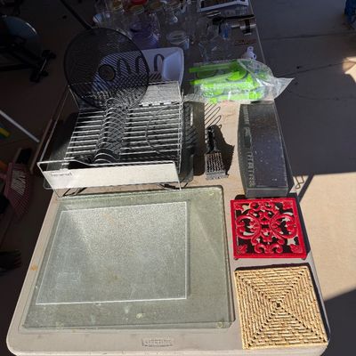 Sale Photo Thumbnail #242: Assorted kitchenware and household items, including a wire dish drying rack, glass cutting boards, trivets, and glassware, displayed on a folding table.