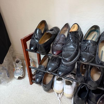 Sale Photo Thumbnail #125: A collection of men's dress shoes, including black leather loafers and wingtips, arranged on a multi-tier wooden and metal shoe rack alongside white Reebok sneakers.