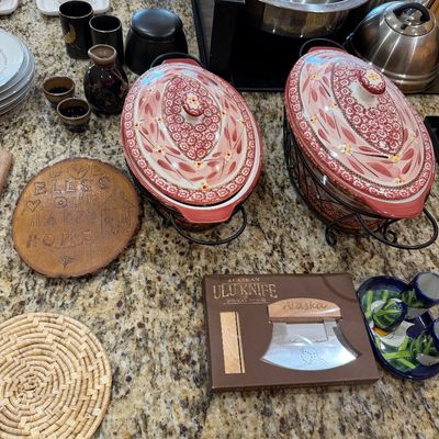 Sale Photo Thumbnail #202: Temp-tations floral ceramic covered casserole dishes in wire stands, shown alongside an Alaskan Ulu knife set, a wood-burned plaque, and various kitchenware items.