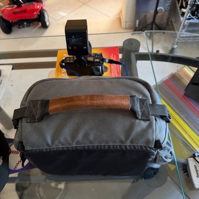 Peak Design grey canvas camera bag with a brown leather handle grip, shown resting on a glass table.