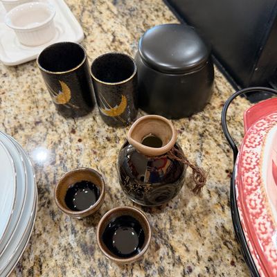 Sale Photo Thumbnail #205: Japanese ceramic sake set featuring a dark brown glazed tokkuri bottle with gold calligraphy and two matching ochoko cups, alongside a pair of black tumblers adorned with golden crane motifs.