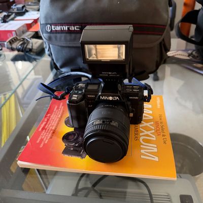 Vintage Minolta Maxxum 7000 35mm SLR camera with attached lens and Maxxum 2800 AF flash, resting on a Minolta Maxxum guidebook with a...