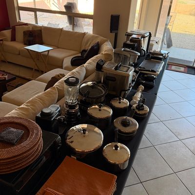 Assorted kitchenware and small appliances, including stainless steel pots, a blender, a slow cooker, and woven chargers, displayed on a...
