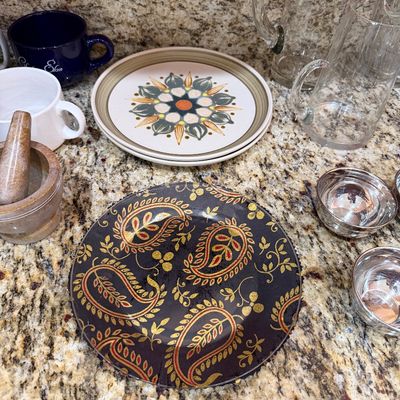 Sale Photo Thumbnail #214: Assorted kitchenware including a black and gold paisley plate, retro floral patterned plates, a stone mortar and pestle, glass pitchers, and small metal bowls.