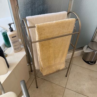Sale Photo Thumbnail #138: Freestanding metal towel rack with a brushed nickel finish, featuring three hanging bars and a bottom shelf, shown holding two bath towels.