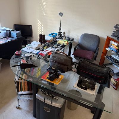 L-shaped glass-top desk with black metal legs, featuring an assortment of electronics, camera gear, office supplies, and a paper shredder...