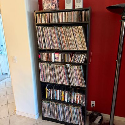 Black laminate media shelving unit filled with a large collection of vinyl records, CDs, and DVDs, topped with a boxed set and small...