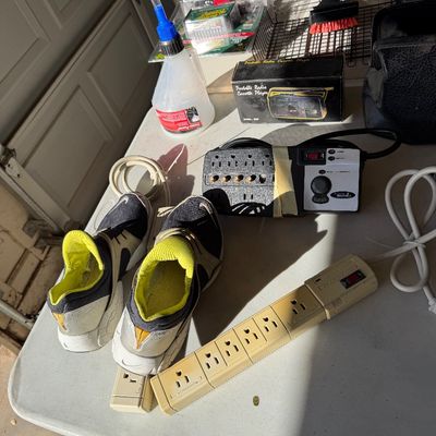 Assorted household items on a folding table, including a pair of Nike athletic shoes, power strips, a portable radio cassette player in...