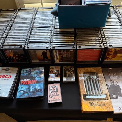 Large collection of CDs in black plastic storage racks, accompanied by a blue zippered media bag, cassette tapes, and DVDs including...