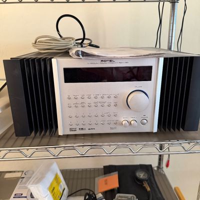 Sale Photo Thumbnail #154: Rotel RSX-1055 surround sound receiver featuring a silver faceplate, prominent volume dial, and large black side heatsinks, displayed on a wire shelf.