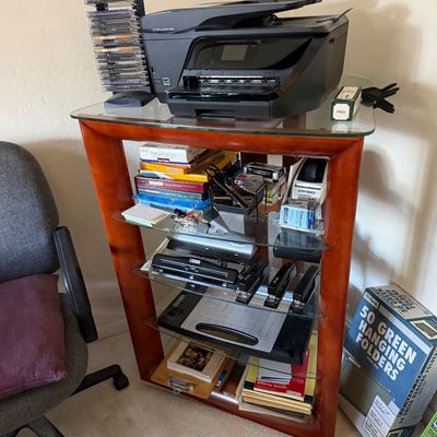 Modern wood-framed shelving unit with glass shelves, holding an HP OfficeJet printer, office supplies, and stationery.
