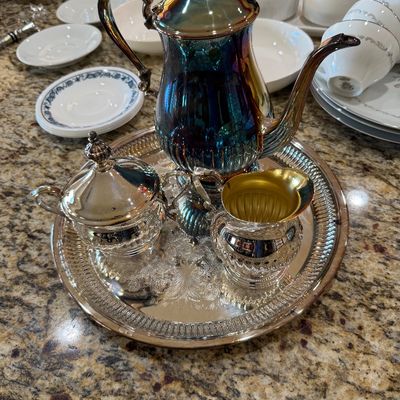 Sale Photo Thumbnail #198: Vintage silver-plated tea service set on an engraved round tray, featuring a heavily tarnished teapot with iridescent blue and gold patina, a lidded sugar bowl, and a creamer with a gold-washed interior.