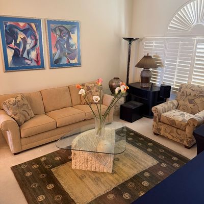 Living room furniture set including a beige three-cushion sofa, a patterned armchair, a triangular glass-top coffee table with a stone...