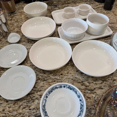 Sale Photo Thumbnail #201: Assortment of white ceramic tableware, including bowls, saucers, ramekins, and cappuccino mugs with metal stands, displayed alongside a blue-patterned saucer.