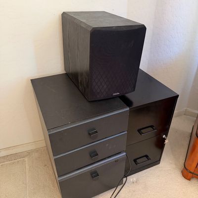 Sale Photo Thumbnail #144: Black wood-grain speaker cabinet resting on a three-drawer mobile pedestal file cabinet next to a two-drawer metal filing cabinet.