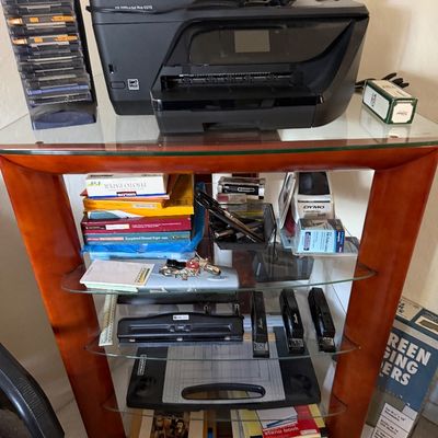 HP OfficeJet Pro 6978 printer sitting atop a wood-framed glass shelving unit filled with office supplies, including staplers, paper, and...