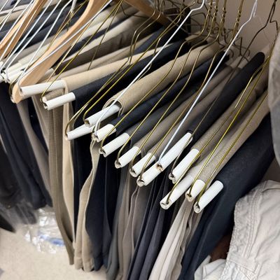 Sale Photo Thumbnail #121: Selection of men's dress trousers in neutral tones, neatly hung on wire and wooden hangers in a closet.