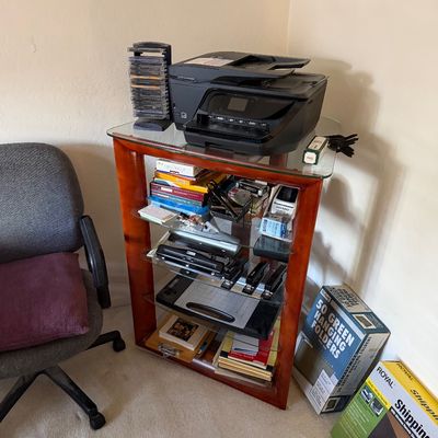 Modern wood and glass shelving unit holding an HP printer, office supplies, and a CD rack, shown next to an office chair and shipping boxes.