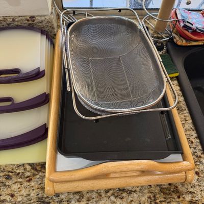 Sale Photo Thumbnail #225: Assorted kitchenware including an over-the-sink stainless steel mesh colander, a black baking sheet, a wooden serving tray, and a stack of plastic cutting boards with purple handles.