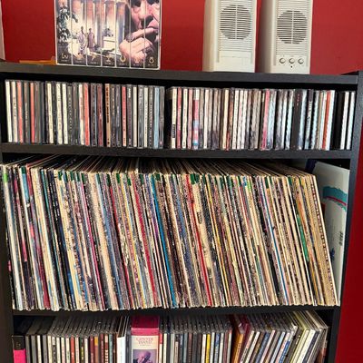 Sale Photo Thumbnail #297: Black wooden media shelf filled with a large collection of vinyl records and CDs, topped with an "I, Claudius" VHS box set and a pair of vintage white computer speakers.