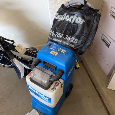 Sale Photo Thumbnail #116: Rug Doctor Mighty Pro carpet cleaning machine, featuring a blue and white plastic body, rolling wheels, and an attached mesh storage bag containing hoses and attachments.