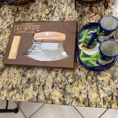 Sale Photo Thumbnail #197: Alaskan Ulu knife and display stand in its original packaging, alongside a hand-painted ceramic condiment set with two small cups and a shaker on a matching tray.