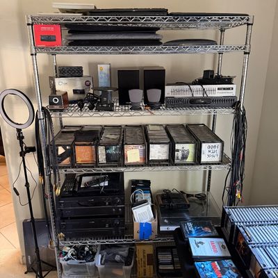 Five-tier wire shelving unit on casters, holding various electronics, audio equipment, CD storage racks, and a ring light on a tripod stand.