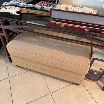 Sale Photo Thumbnail #288: Upholstered beige storage ottoman tucked beneath a glass-top console table, shown alongside a poker chip set, electric guitar, and vintage computer tower.