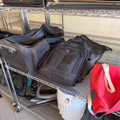 Assorted travel bags, backpacks, and luggage displayed on wire shelving units, including a red canvas tote and a white Igloo water cooler.