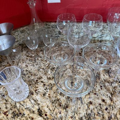 Sale Photo Thumbnail #215: Assorted glassware collection including a glass decanter with stopper, coupe glasses, wine glasses, and a small crystal vase, displayed on a granite countertop.