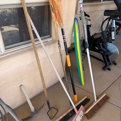 Sale Photo Thumbnail #237: Assorted outdoor tools including a pickaxe, garden hoe, push brooms, a leaf rake, and extension poles, showing signs of regular use.