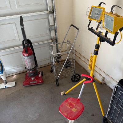 Sale Photo Thumbnail #106: Assorted household and utility items, including a Hoover upright vacuum, a folding wire shopping cart, a yellow dual-head halogen work light on a tripod, and a red 1999 Phoenix Open folding spectator seat.