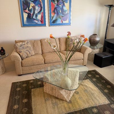 Sale Photo Thumbnail #316: Contemporary living room set featuring a beige upholstered sofa, a triangular glass-top coffee table with a textured stone base, and a pair of framed abstract art prints.