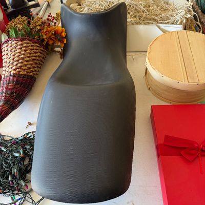 Molded black vinyl motorcycle seat, showing light wear, displayed on a table with various household and holiday items.