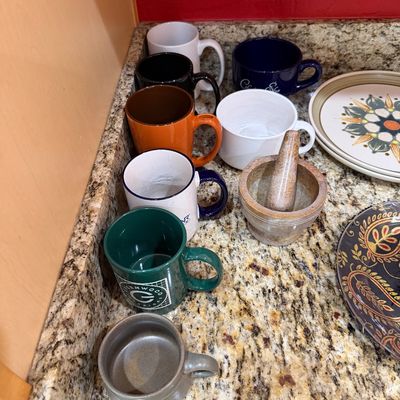 Sale Photo Thumbnail #218: Assorted ceramic coffee mugs in various colors, a stone mortar and pestle, and patterned plates displayed on a granite countertop.