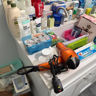Sale Photo Thumbnail #90: Assorted bathroom toiletries and personal care items, including an orange hair dryer, Aveeno lotion, Cetaphil cream, and Crest toothpaste, arranged on a vanity countertop.