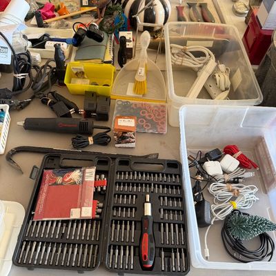 Sale Photo Thumbnail #107: Assorted tools and electronics accessories, including a multi-bit screwdriver set, a heat gun, a crowbar, and various power cords and chargers organized on a table.